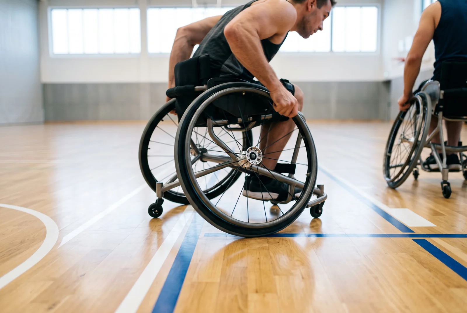 Two athletes in sports wheelchairs moving across a polished hardwood indoor court floor.